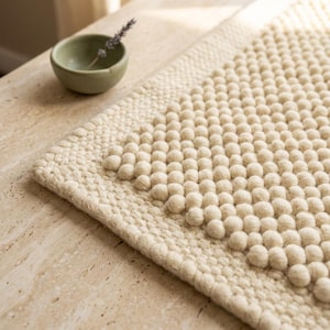 Puede incluir: Alfombra texturizada de color crema con un patrón de acentos esféricos en relieve. La alfombra tiene un borde grueso y tejido y una apariencia suave y acogedora. Un pequeño cuenco verde con una ramita de lavanda está cerca.