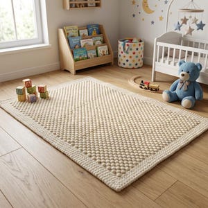 May include: Cream-coloured rectangular rug with a textured, dotted pattern. The rug is on a wooden floor in a nursery, with a cot, teddy bear, and wooden toy blocks visible. The rug measures approximately 150cm x 90cm.