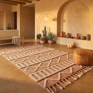 May include: A rectangular rug with a geometric pattern in tan and cream. The rug has a fringe along the bottom edge and is placed on a light brown floor. The room also features cacti, pottery, and a leather ottoman. The overall style is bohemian.