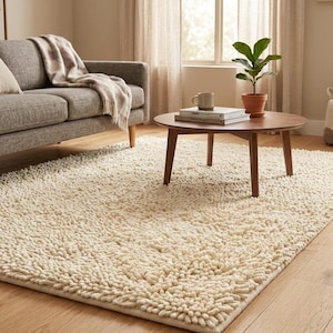 Puede incluir: Una alfombra cremosa y peluda en un salón. La alfombra rectangular cubre una gran parte del suelo de madera. También se ven un sofá gris, una mesa de centro redonda de madera y una planta en maceta.