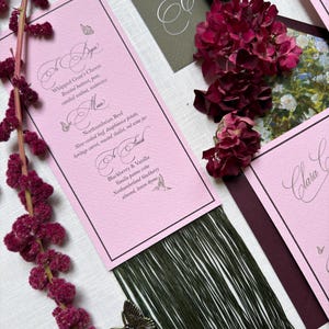May include: A rectangular pink menu card with black text, featuring sections titled To Begin, Main, and To Finish. The card has a dark green fringe at the bottom, a metal butterfly charm, and is surrounded by dark red flowers and green stationery.