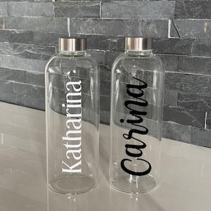 Könnte beinhalten: Zwei transparente Glaswasserflaschen mit silbernen Deckeln. Eine Flasche trägt den Namen "Katharina" in weißer Schrift, die andere "Carina" in schwarzer Schrift. Die Flaschen stehen auf einer hellen Oberfläche vor einer dunkelgrauen Steinmauer.