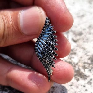 May include: A silver-toned wing-shaped pendant with black accents. The pendant features a detailed feather design, with small black stones clustered along the edges. The wing is held in a hand against a blurred background.