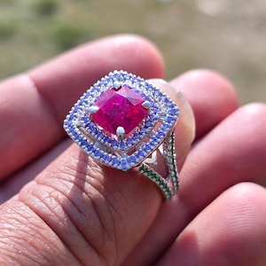 May include: A square-cut ruby ring with a double halo of blue and clear stones. The ring is held in a hand, with the brand name "Ankit Diamond Jewels" visible in the background.