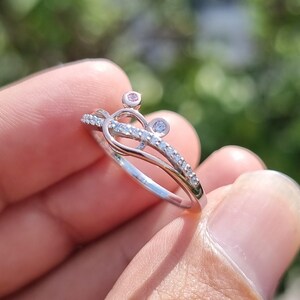 May include: A silver ring with a crown design, featuring small clear and pink gemstones. The ring is held in a hand against a blurred green background. The text "Ankit Diamond Jewels" is visible at the bottom of the image.