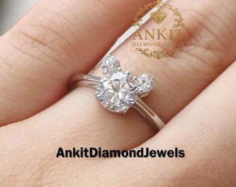 Könnte beinhalten: Ein silberner Ring mit einem großen, runden, klaren Diamanten in der Mitte und zwei kleineren Diamanten auf jeder Seite, die eine Mickey-Mouse-Form bilden. Der Ring befindet sich an einem Finger. Der Text "AnkitDiamondJewels" steht unten.