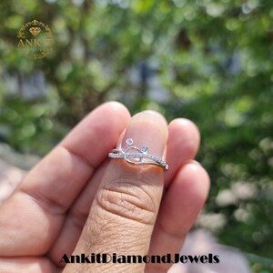 May include: A silver ring with a unique design, featuring small clear gemstones and a delicate, curved band. The ring has a small, circular design with two accent stones. The ring is held up against a blurred green background.