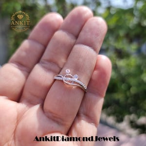 May include: A silver ring with a unique design featuring small, round diamonds. The ring has a delicate band with additional small diamonds. The ring is held up by a hand. The text "Ankit Diamond Jewels" is at the bottom.