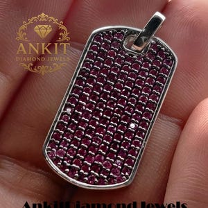 Puede incluir: Colgante rectangular con marco plateado, densamente engastado con pequeñas gemas redondas de color rojo intenso. El colgante tiene un lazo en la parte superior para una cadena. El texto "Ankit Diamond Jewels" está en la parte inferior.