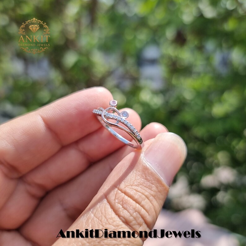 May include: A silver ring with a crown design, featuring small clear and pink gemstones. The ring is held in a hand against a blurred green background. The text "Ankit Diamond Jewels" is visible at the bottom of the image.