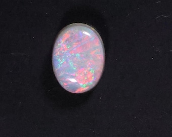 May include: An oval-shaped opal gemstone with a white base and iridescent flashes of pink, red, green, and blue. The polished surface reflects light, showcasing the stone's internal play of color. The gemstone is set against a black background.