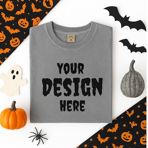 May include: A gray t-shirt with the text "YOUR DESIGN HERE" in black, surrounded by Halloween decorations. Includes a pumpkin, bats, a ghost, and spider. The shirt is a blank canvas for custom designs.
