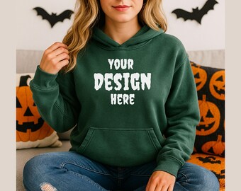 May include: A dark green hoodie with the text "YOUR DESIGN HERE" in white. The hoodie has a front pocket and a hood. The person is wearing blue jeans. Halloween-themed pillows and bat decorations are in the background.
