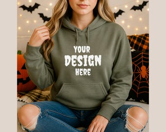 May include: Olive green hoodie with the text "YOUR DESIGN HERE" in white. The hoodie has a front pocket and a drawstring hood. The model is wearing blue jeans. Halloween decorations are in the background.