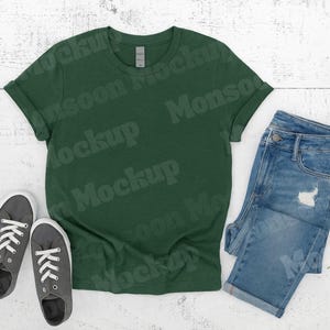 Peut inclure: Un t-shirt vert foncé à manches retroussées, associé à un jean en denim bleu et des baskets grises. Les articles sont disposés sur une surface en bois blanc, créant un look décontracté et quotidien.
