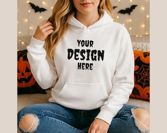 May include: White hoodie with the text "YOUR DESIGN HERE" in a black, distressed font. The hoodie has a front pocket and a drawstring hood. The model is wearing blue jeans. Halloween-themed decor is in the background.