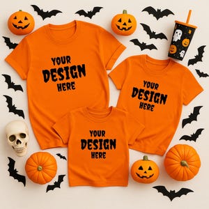 May include: Orange t-shirts with the text "YOUR DESIGN HERE" are displayed with Halloween decorations. The decorations include pumpkins, bats, a skull, and a Halloween-themed cup. The shirts are in various sizes.
