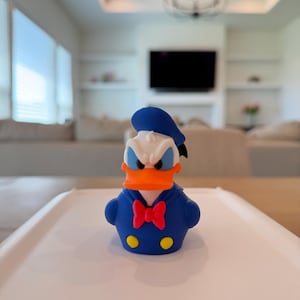 May include: A blue and white Donald Duck figurine with an orange beak and a red bow tie. The figurine is wearing a blue sailor suit and hat, and has yellow buttons on the front.