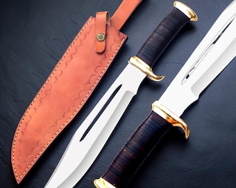 May include: A fixed-blade knife with a silver steel blade featuring a long fuller, a dark brown handle, and a gold-colored guard and pommel. Beside the knife is a tan leather sheath with a snap closure and decorative stitching along the edges.
