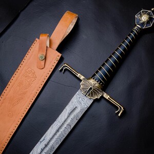 Op de afbeelding: Een zwaard met een patroon van Damascusstaal, een zwart handvat met gouddraadwikkeling en een messing stootplaat en pommel. De pommel heeft een zonmotief. Naast het zwaard ligt een lichtbruine lederen schede met bloemmotieven.