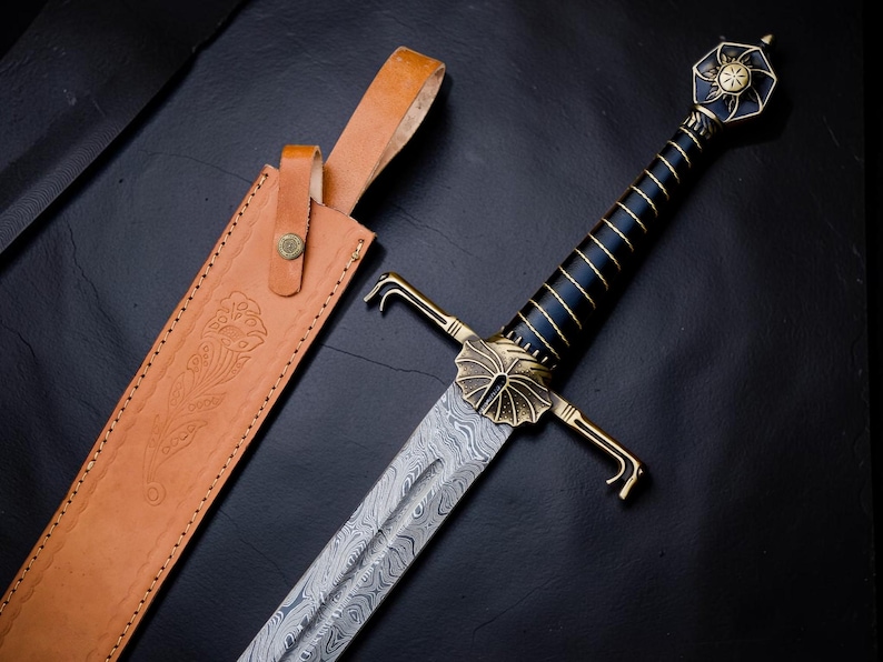 Op de afbeelding: Een zwaard met een patroon van Damascusstaal, een zwart handvat met gouddraadwikkeling en een messing stootplaat en pommel. De pommel heeft een zonmotief. Naast het zwaard ligt een lichtbruine lederen schede met bloemmotieven.