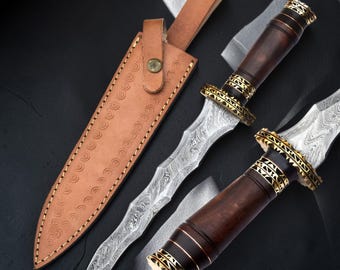 May include: A Damascus steel dagger with a wavy blade, a dark wood handle, and gold-colored metal accents. The dagger is accompanied by a tan leather sheath featuring a stamped circular pattern and a small metal snap button closure.