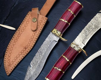 May include: A fixed-blade knife with a patterned Damascus steel blade and a deep red handle featuring gold-colored metal bands. The knife is accompanied by a tan leather sheath with decorative stitching and a single brass-colored snap button.