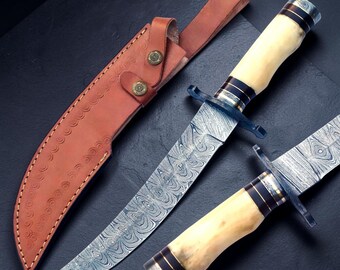 May include: Two fixed-blade knives with patterned Damascus steel blades and cream-colored handles featuring dark bands. Each knife has a metal crossguard. A tan leather sheath with decorative stitching and two brass-colored metal snaps is included.