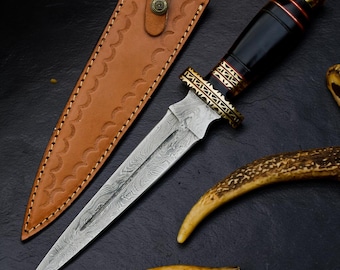 May include: A fixed-blade knife with a Damascus steel blade featuring a wavy pattern, a black handle with gold-colored metal accents, and a tan leather sheath with decorative stitching and a metal snap button closure.