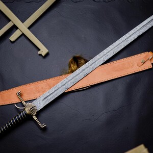 Op de afbeelding: Een lang zwaard met een patroon van Damascusstaal, een zwarte geribbelde greep en een messing stootplaat met een schelpachtig ontwerp. Het zwaard bevat een lichtbruine lederen schede met reli&euml;fdetails en een messing knoopsluiting.