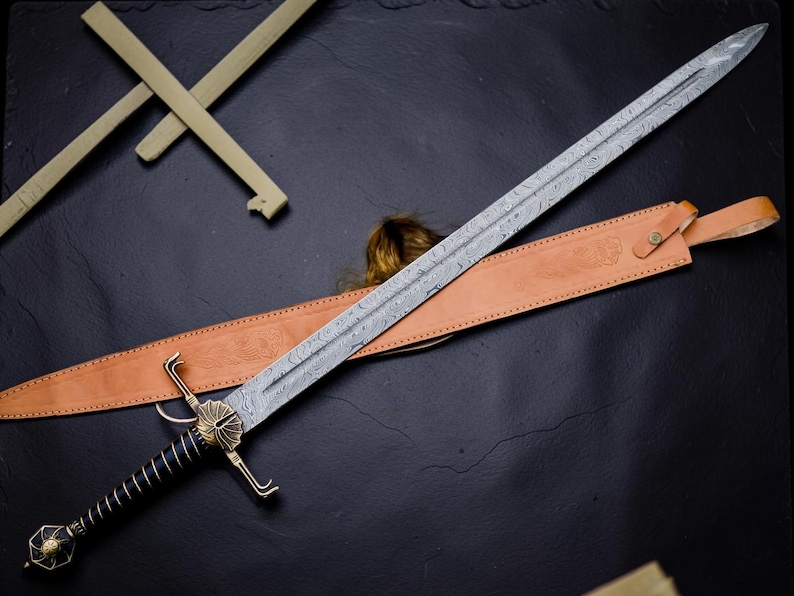 Op de afbeelding: Een lang zwaard met een patroon van Damascusstaal, een zwarte geribbelde greep en een messing stootplaat met een schelpachtig ontwerp. Het zwaard bevat een lichtbruine lederen schede met reli&euml;fdetails en een messing knoopsluiting.