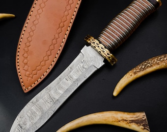 May include: A fixed-blade knife with a Damascus steel blade featuring a wavy pattern, a handle wrapped in alternating silver and copper wire, and a decorative brass guard. It is accompanied by a tan leather sheath with stitched edges.