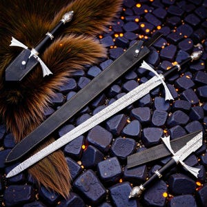 May include: A collection of medieval-style swords with ornate silver crossguards and black handles. The blades have a detailed pattern, and the swords are displayed with a black leather sheath and a matching dagger. The background features dark blue stones and faux fur.