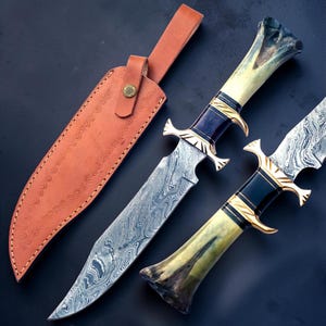 May include: Two fixed-blade knives with Damascus steel blades featuring a wavy pattern, gold-colored metal guards, and cream-colored bone handles with dark accents. A tan leather sheath with embossed spiral patterns and a brass snap button is included.