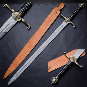 Op de afbeelding: Een lang zwaard met een patroon van Damascusstaal, een zwart handvat met gouden draadwikkeling en een messing stootplaat met een waaiervormig ontwerp. Het zwaard wordt vergezeld door een beige lederen schede met reli&euml;f bloemmotieven.