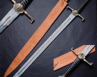 Op de afbeelding: Een lang zwaard met een patroon van Damascusstaal, een zwart handvat met gouden draadwikkeling en een messing stootplaat met een waaiervormig ontwerp. Het zwaard wordt vergezeld door een beige lederen schede met reliëf bloemmotieven.