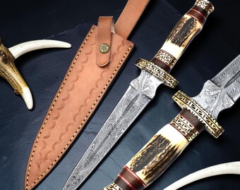 May include: Two double-edged daggers with Damascus steel blades, handles made of textured antler and dark wood with gold-toned metal accents, and a tan leather sheath featuring a snap button closure and decorative crescent-shaped stitching.