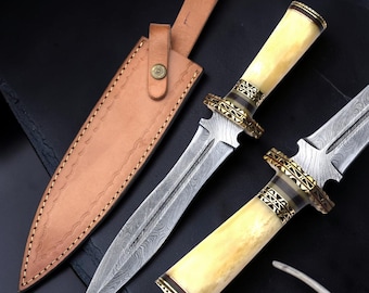 Op de afbeelding: Een tweesnijdende dolk met een patroon van Damascusstaal, een crèmekleurig handvat en decoratieve goudkleurige metalen accenten. De dolk wordt vergezeld door een beige lederen schede met een drukknoopsluiting en gestikte randdetails.