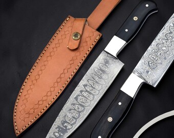 Op de afbeelding: Twee messen met een patroon van Damascusstaal en zwarte handvatten met koperen klinknagels, vergezeld van een lichtbruine lederen schede met decoratieve stiksels en een metalen drukknoop. De bladen hebben een opvallende golvende metaalstructuur.