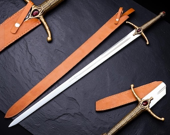 May include: A long sword with a silver blade and a textured brass-toned hilt featuring a red oval gemstone. The sword is accompanied by a tan leather scabbard with a metal snap closure. Two additional sword hilts with matching red gemstones are shown.