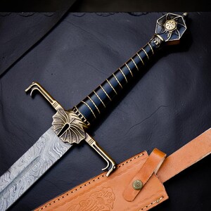 Op de afbeelding: Een zwaard met een Damascus stalen lemmet met een golvend patroon, een zwarte greep met gouddraadwikkeling en een messing pareerstang in de vorm van een waaier. De pommel is zeshoekig met een zonnewijzerontwerp. Een beige lederen schede is inbegrepen.