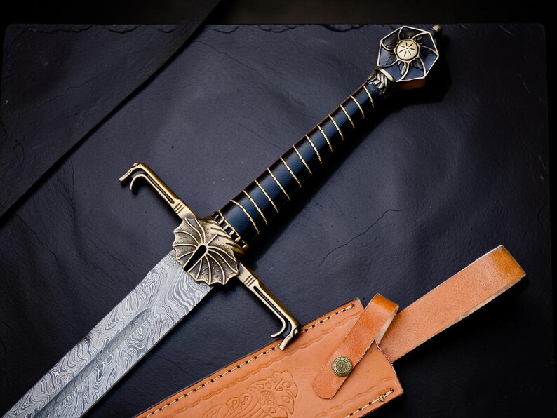 Op de afbeelding: Een zwaard met een Damascus stalen lemmet met een golvend patroon, een zwarte greep met gouddraadwikkeling en een messing pareerstang in de vorm van een waaier. De pommel is zeshoekig met een zonnewijzerontwerp. Een beige lederen schede is inbegrepen.