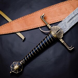 Op de afbeelding: Een zwaard met een patroon van Damascusstaal, een zwart handvat met goudkleurig draadwikkelwerk, en een messingkleurige stootplaat en pommel met bloemmotieven. Het zwaard wordt vergezeld door een beige lederen schede met bladreli&euml;fs.