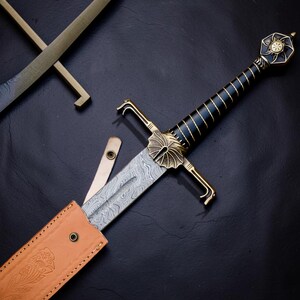 Op de afbeelding: Een zwaard met een patroon van Damascusstaal, een zwart handvat met gouden draadomwikkeling en een goudkleurige pareerstang met een waaiervormig ontwerp. De pommel heeft de vorm van een zon. Het zit gedeeltelijk in een beige lederen schede.