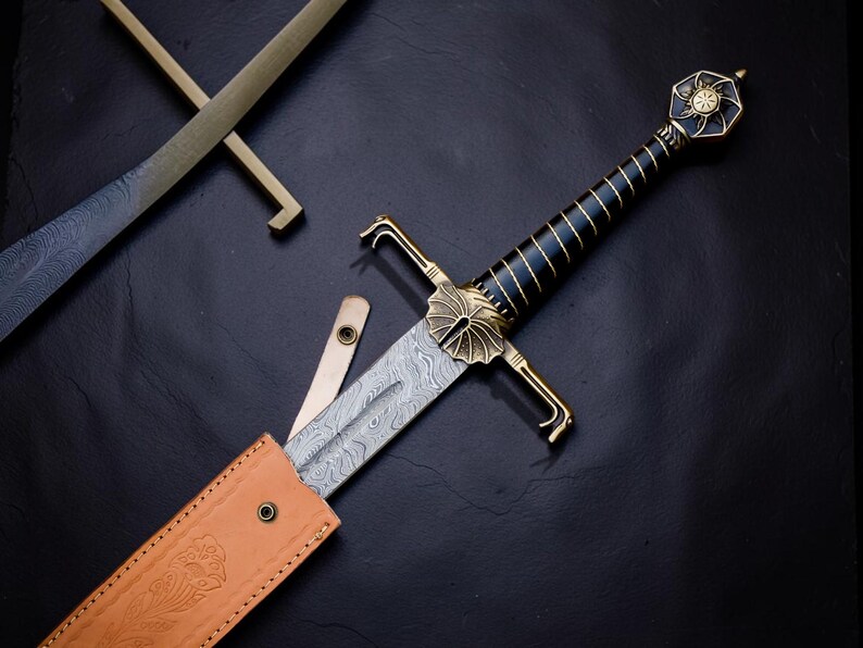 Op de afbeelding: Een zwaard met een patroon van Damascusstaal, een zwart handvat met gouden draadomwikkeling en een goudkleurige pareerstang met een waaiervormig ontwerp. De pommel heeft de vorm van een zon. Het zit gedeeltelijk in een beige lederen schede.