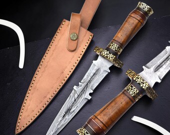 May include: A double-edged dagger with a patterned Damascus steel blade, a brown wooden handle, and gold-colored metal accents. The dagger is accompanied by a tan leather sheath featuring a snap button closure and decorative stitching along the edges.