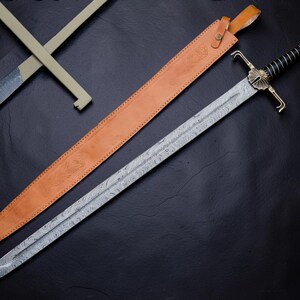 Op de afbeelding: Een lang zwaard met een patroon van Damascus staal, een zwart omwikkeld handvat en een messing stootplaat. Het zwaard heeft een decoratieve pommel en wordt vergezeld door een beige lederen schede met een riem en metalen gespsluiting.