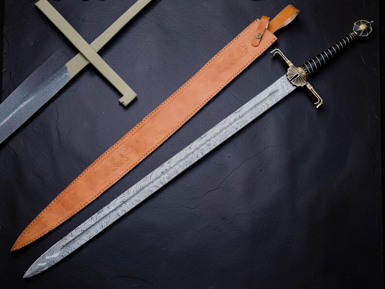 Op de afbeelding: Een lang zwaard met een patroon van Damascus staal, een zwart omwikkeld handvat en een messing stootplaat. Het zwaard heeft een decoratieve pommel en wordt vergezeld door een beige lederen schede met een riem en metalen gespsluiting.