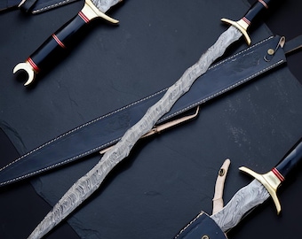 Op de afbeelding: Een zwaard met een golvend lemmet en een Damascus stalen patroon, voorzien van een zwart handvat met rode accenten, een messing stootplaat en een halvemaanvormige pommel. Het zwaard wordt vergezeld door een zwarte lederen schede met bloemmotieven.