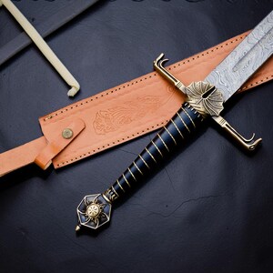 Op de afbeelding: Een zwaard met een patroon van Damascusstaal, een zwart handvat omwikkeld met gouddraad en een messing stootplaat. De pommel heeft een zonnestraalontwerp. Het wordt vergezeld door een beige lederen schede met in reli&euml;f aangebrachte bloemdetails.